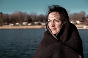 Eine Frau am Meer ist in unsere Decke "Airde" aus Walkloden gekuschelt und guckt in die Sonne.